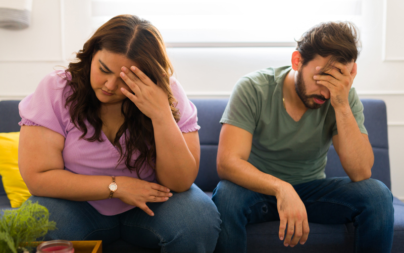 Ein Paar bestehend aus einem Mann und einer übergewichtigen Frau sitzt auf einer Couch und macht eine deprimierten Eindruck.