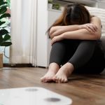 Eine Frau mit depressiver Verstimmung sitzt vor einer Wage und ist unzufrieden mit ihrem Körpergewicht.